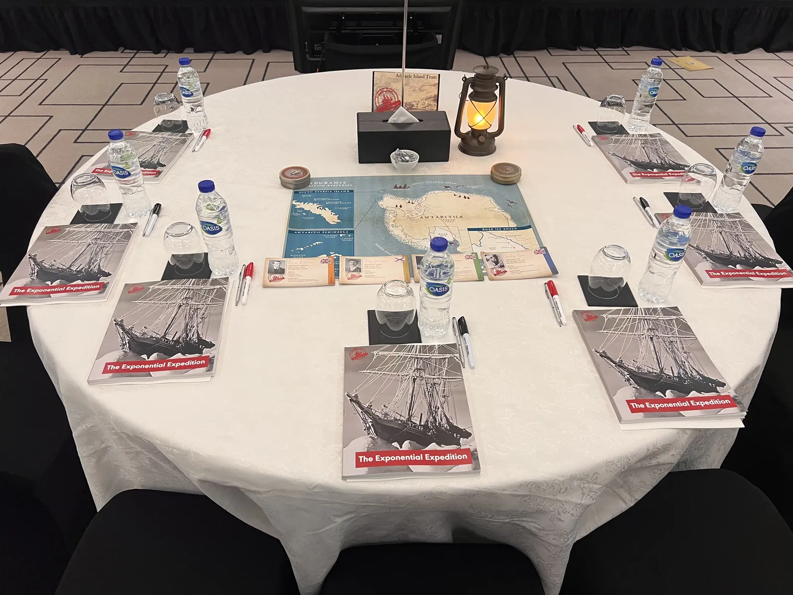 Close-up of a cabaret-style table set with expedition workbooks, an Antarctica route map, a lantern, and crew cards for a Lead the Endurance experience.