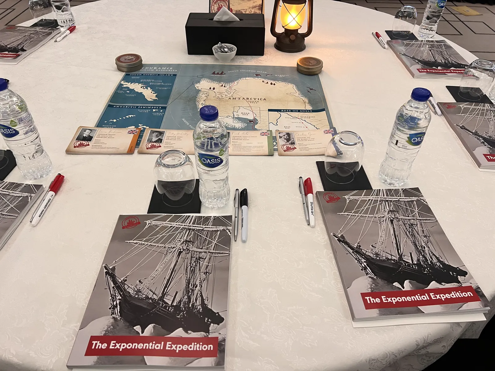 A cabaret-style table set for Lead the Endurance, with expedition workbooks, a route map, a lantern, and pens at every seat.