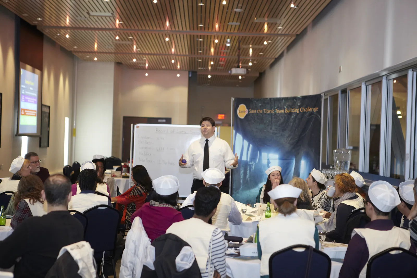 A facilitator leads a debrief with participants seated at cabaret-style round tables during a Save the Titanic team development experience.