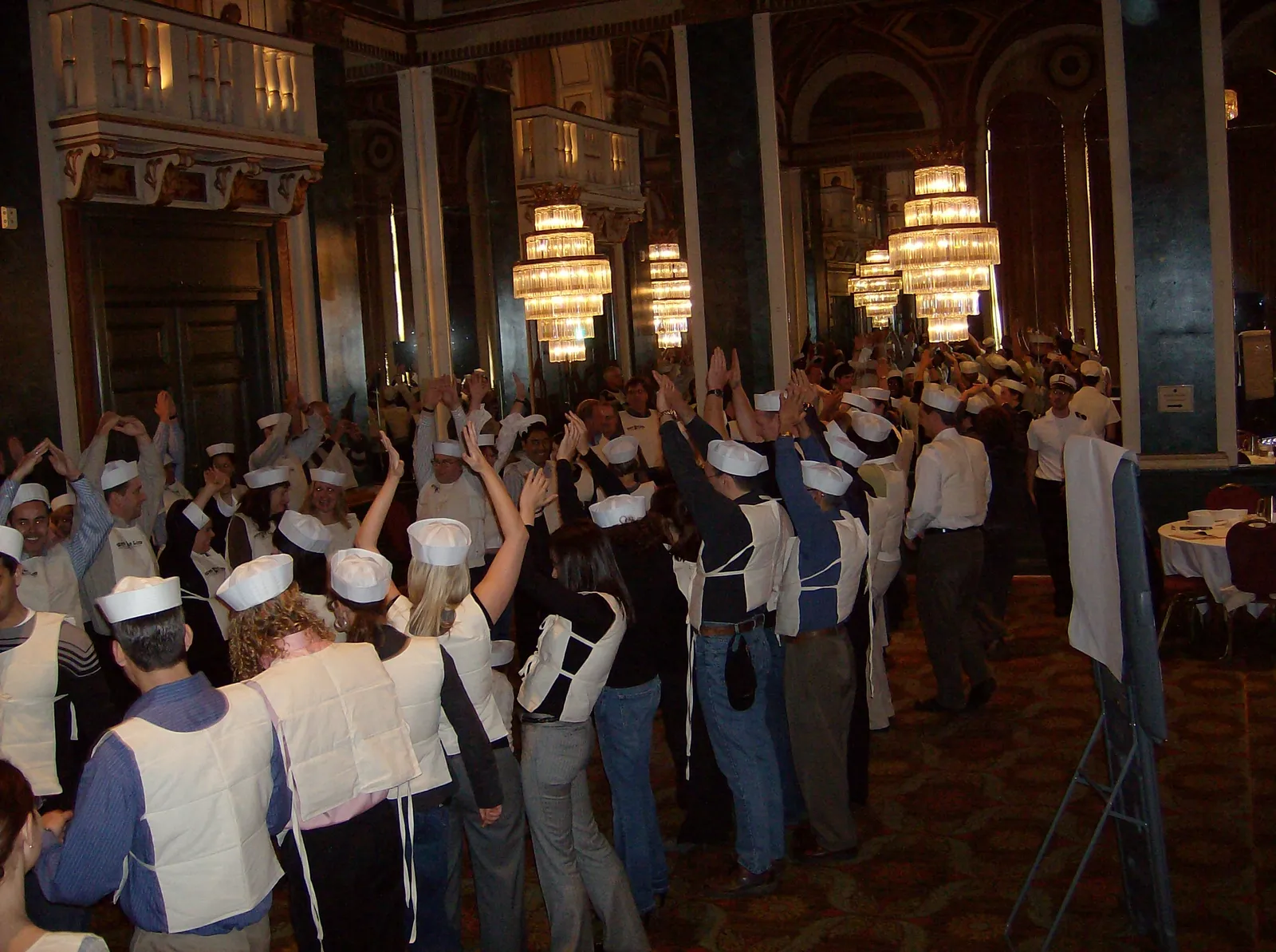 Hundreds of participants stand at cabaret-style tables with hands raised during a large-scale Save the Titanic experience in a grand ballroom.